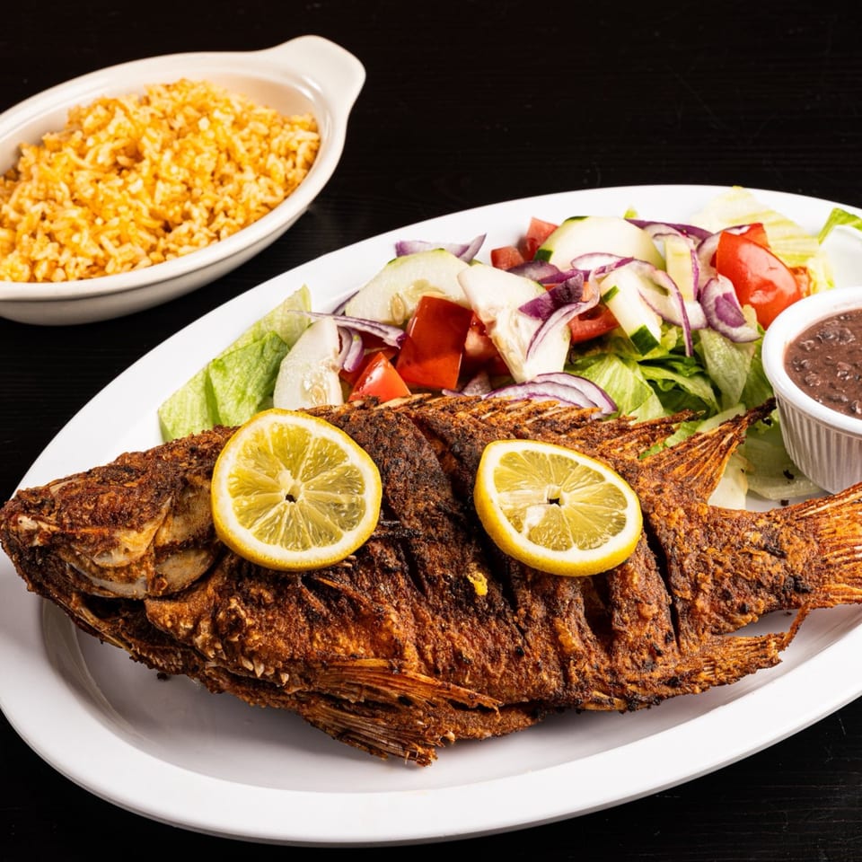 Whole fried fish with lemon slices, side salad, black beans, and rice on a white plate.