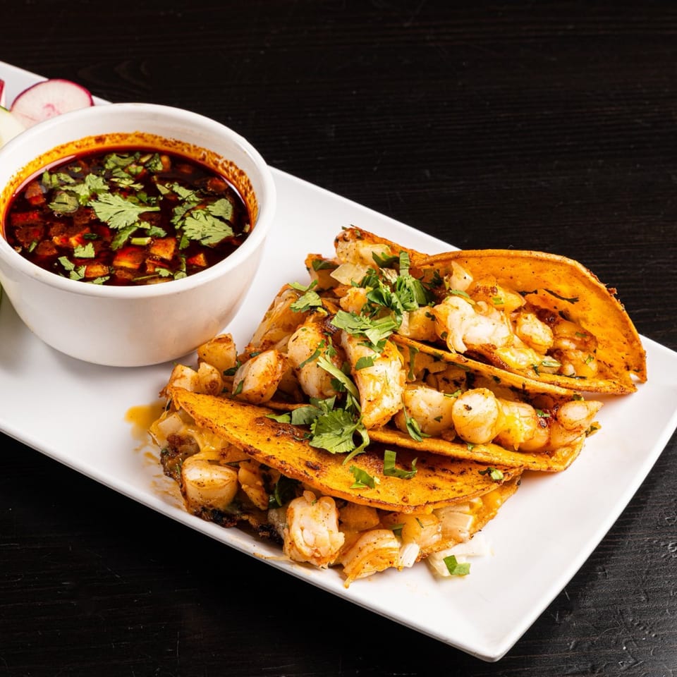 Three shrimp tacos with cilantro and onions, served with a side of red consommé.