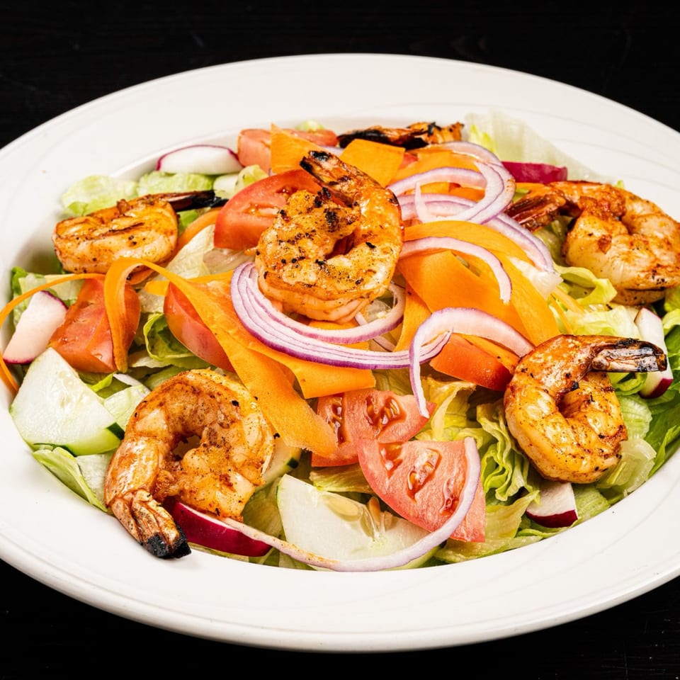 Grilled shrimp salad with lettuce, tomatoes, red onions, carrots, cucumbers, and radishes in a white bowl.