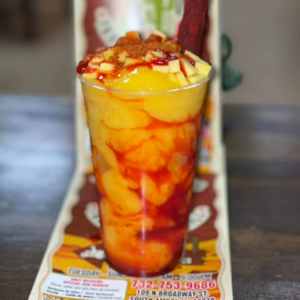 A vibrant Tex-Mex mango drink with chili powder and red sauce, served in a clear plastic cup.