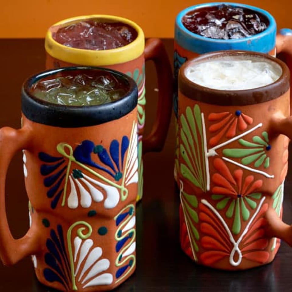 Four colorful, hand-painted Tex-Mex mugs filled with iced drinks on a dark table.