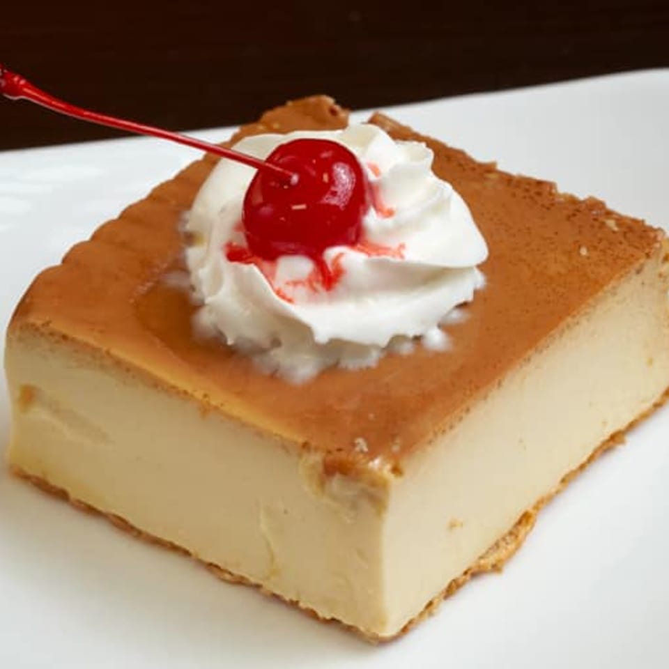 A square slice of flan topped with whipped cream and a maraschino cherry on a white plate.