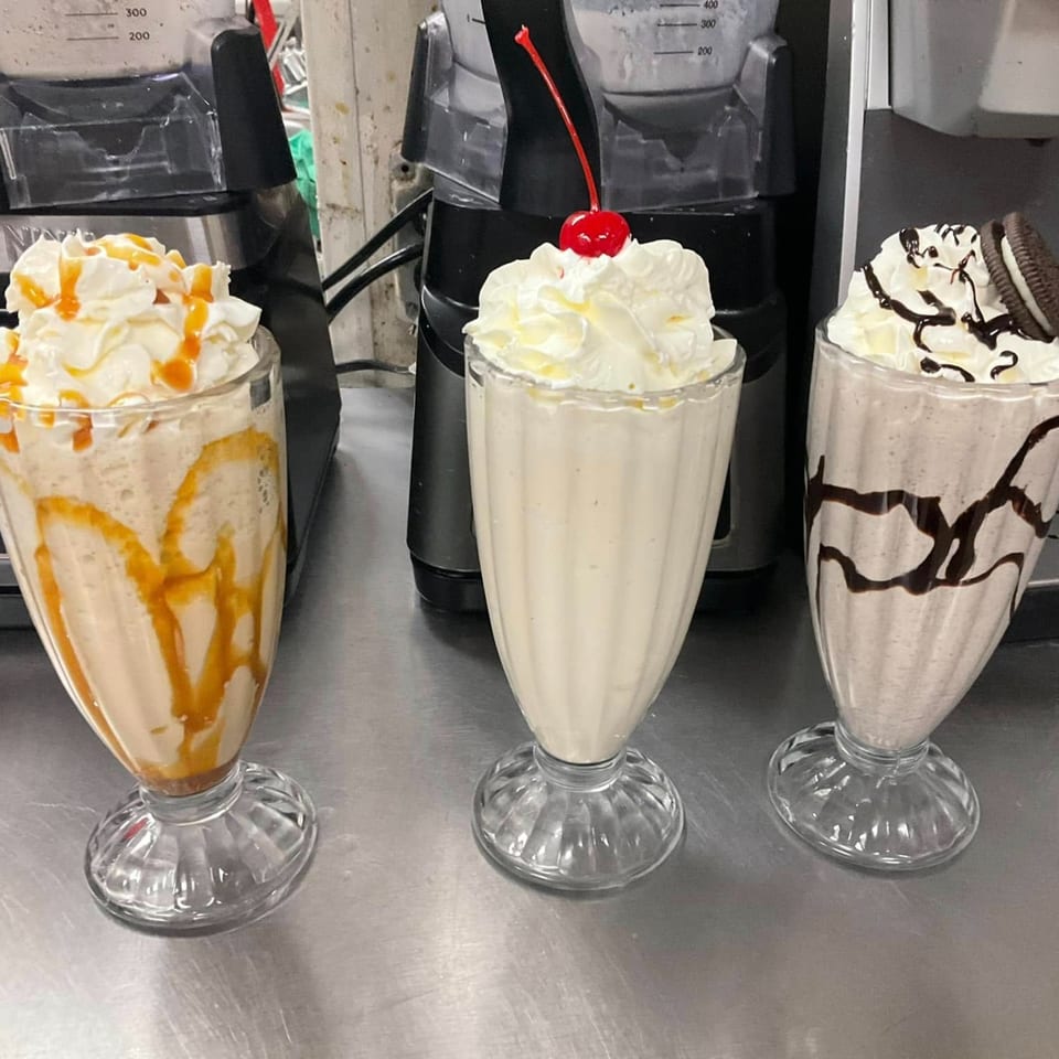 Three milkshakes on a metal counter: caramel, vanilla with a cherry, and Oreo with chocolate drizzle.