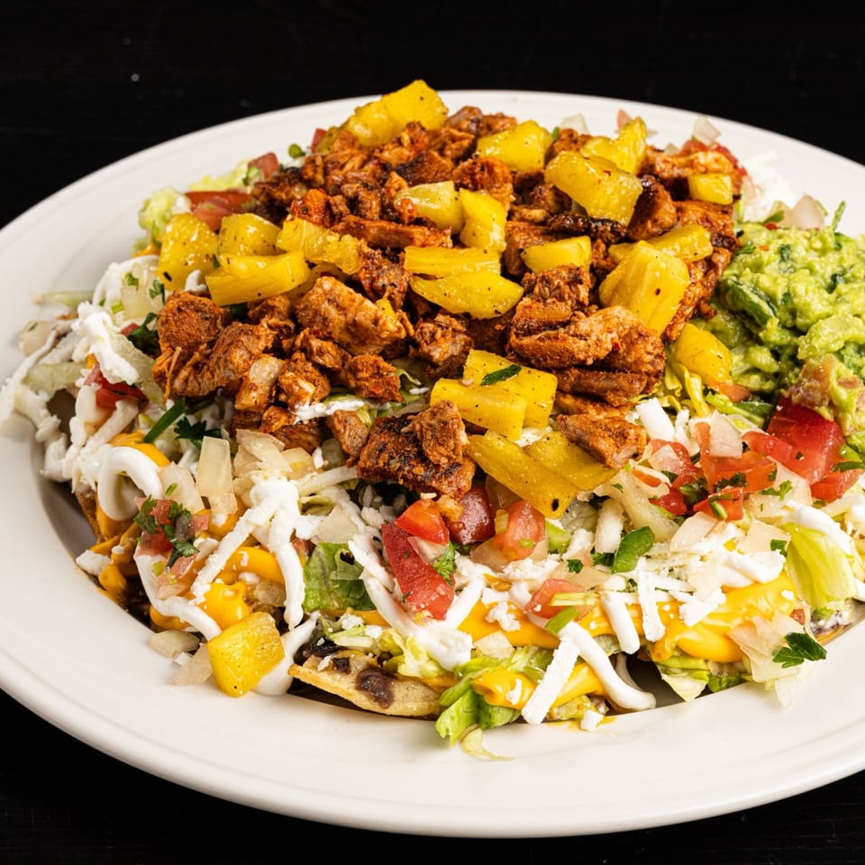 Al pastor nachos with pineapple, guacamole, cheese, and sour cream on a white plate.